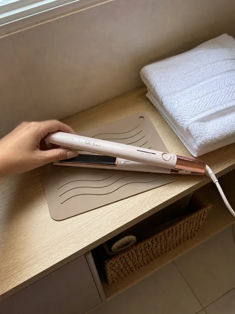 Mão segurando a prancha Britânia BPR04 rosa sobre um tapete de silicone, na prateleira de madeira com toalhas brancas